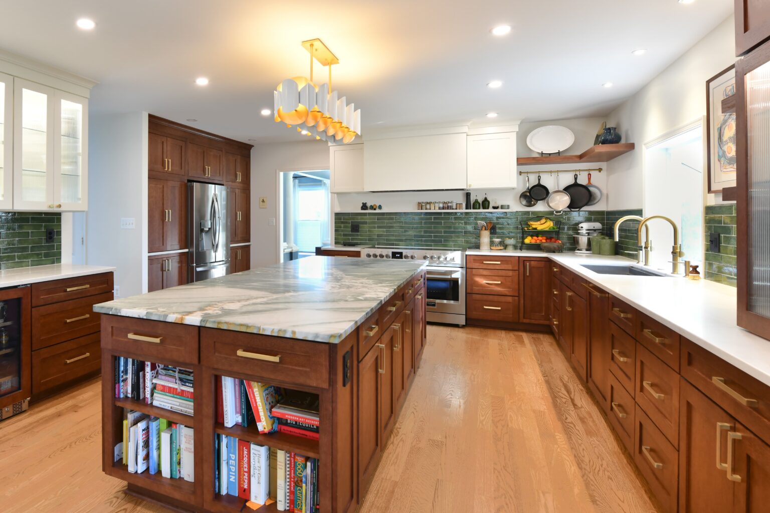 Modern kitchen with wooden cabinets, marble island with built-in bookshelves, green tile backsplash, open shelves, stainless steel appliances, and wood floors under recessed and pendant lighting.