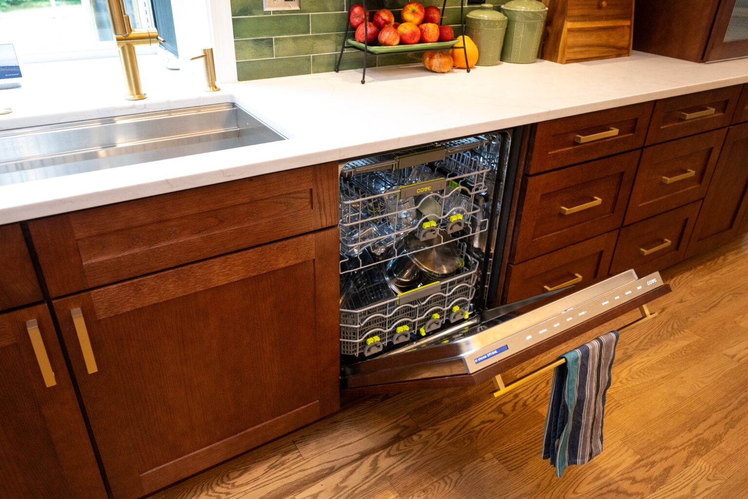 A modern kitchen with an open dishwasher full of dishes, next to a sink and wooden cabinets. A towel hangs on the dishwasher door.