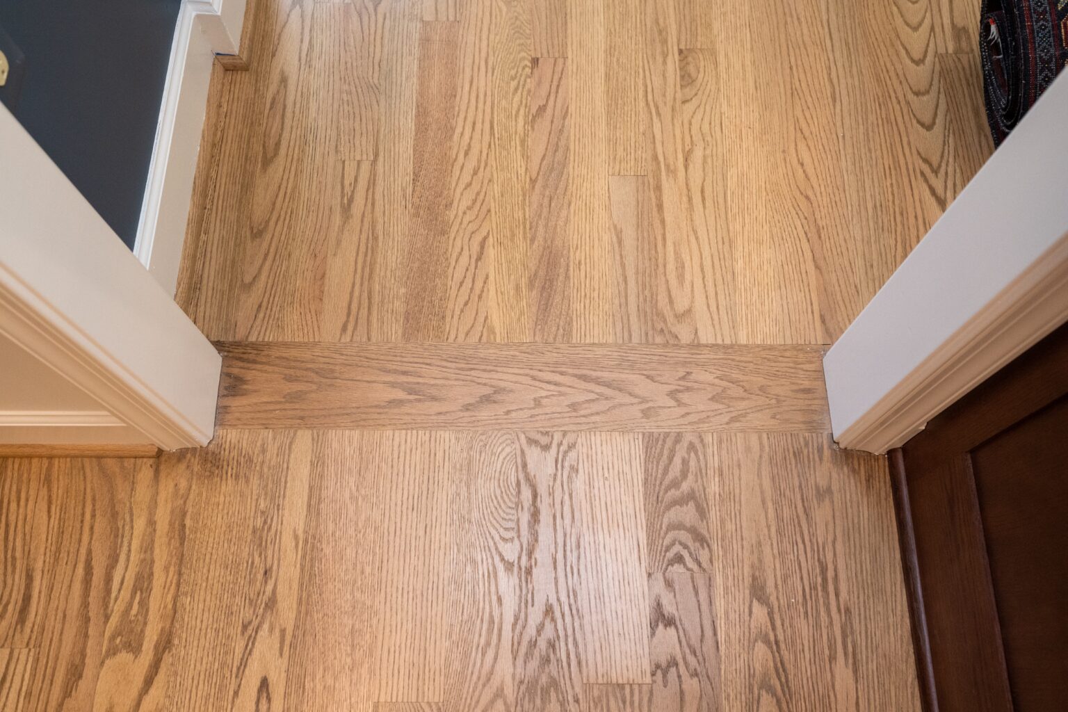 Light-colored hardwood floor with a perpendicular transition board between two doorways, showing varying wood grain patterns.