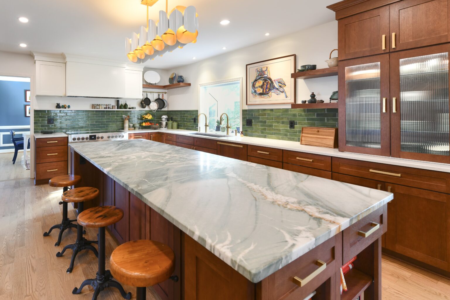 Spacious kitchen with wood cabinets, a marble island countertop, green tile backsplash, bar stools, modern lighting, and decorative shelves.