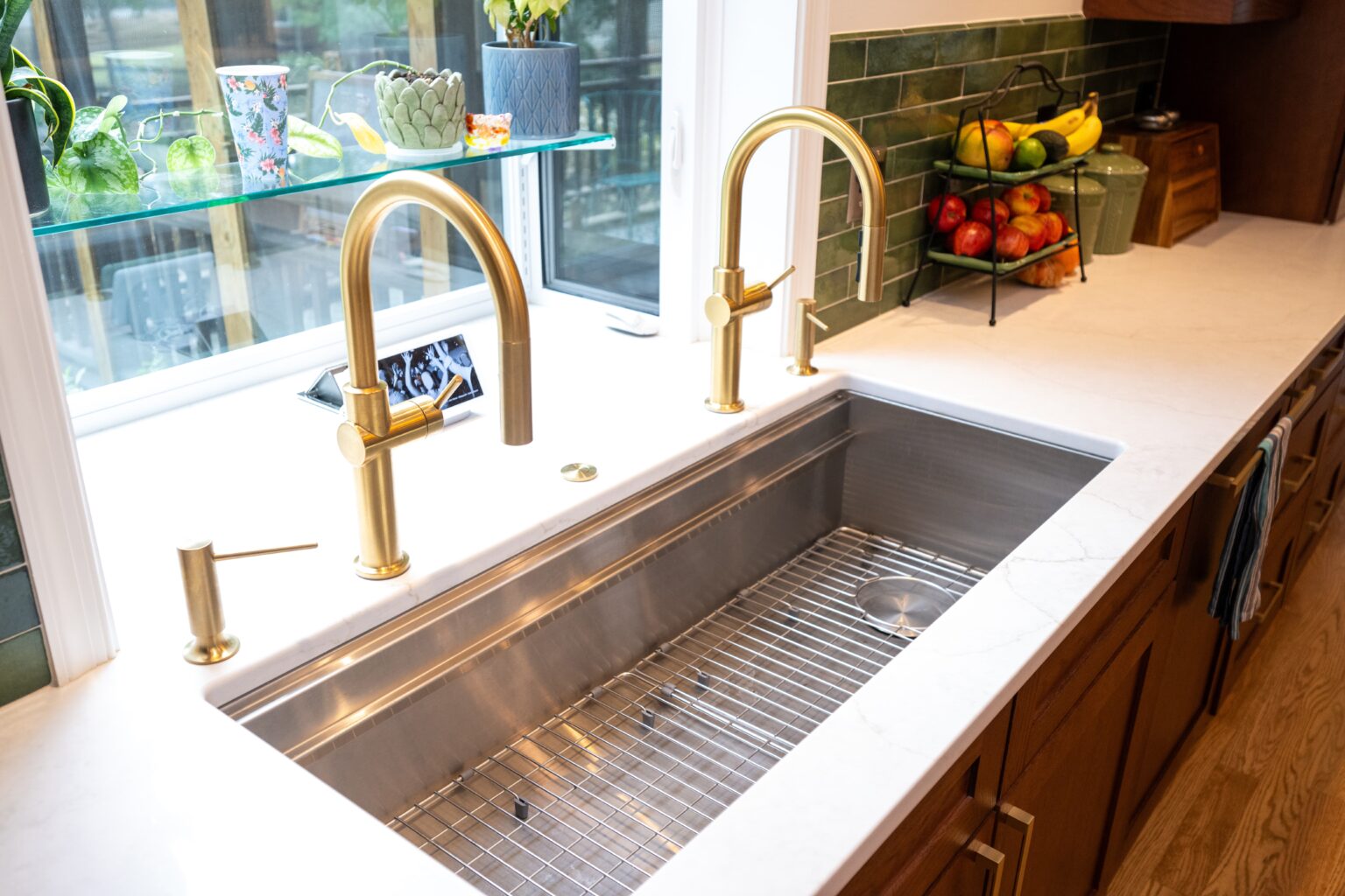 Modern kitchen sink with brass faucets, soap dispenser, and wire rack; fruit bowl and plants on the counter; green tile backsplash; window above the sink.