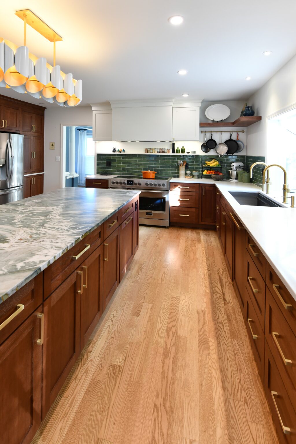Modern kitchen with wooden cabinets, marble and white countertops, stainless steel appliances, green tile backsplash, and open shelving with dishes and cookware. Brightly lit with ceiling lights.