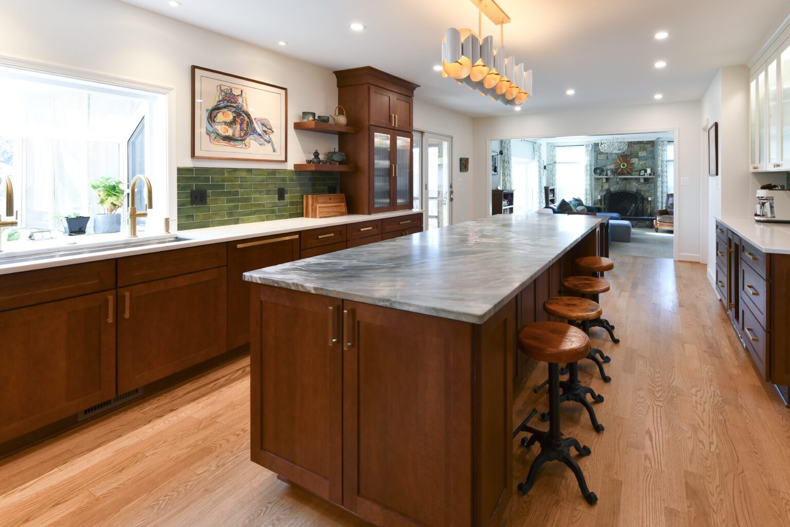 Modern kitchen with a long marble island, wooden cabinets, green tile backsplash, wooden stools, and bright lighting.