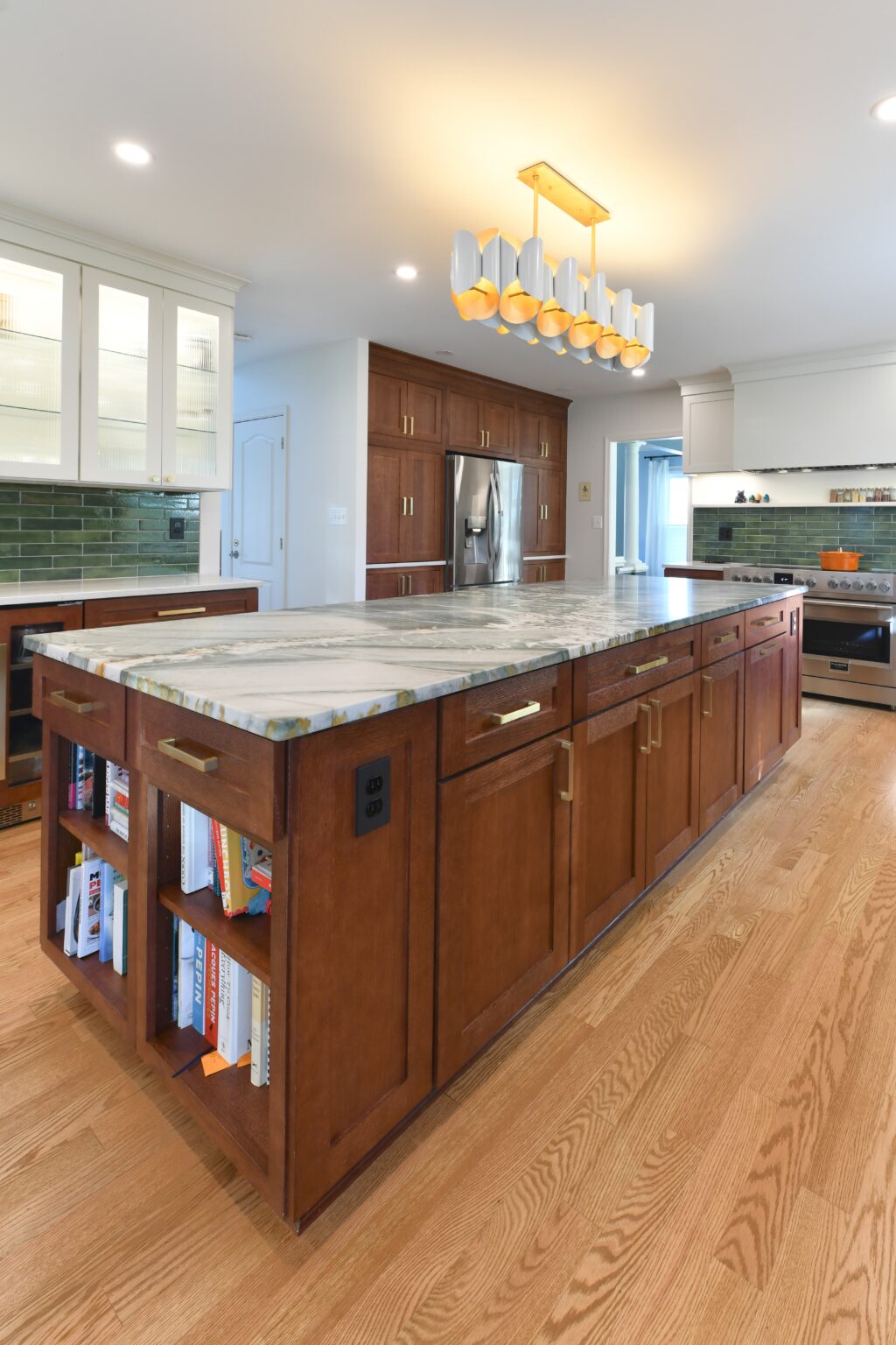 Modern kitchen with a large wooden island featuring a marble countertop, built-in shelves with books, hardwood floors, stainless steel appliances, and contemporary light fixture overhead.