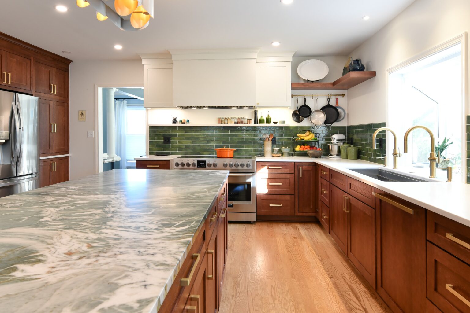 Modern kitchen with wooden cabinets, green tile backsplash, marble island countertop, stainless steel appliances, open shelves, and hanging pots near a window with natural light.