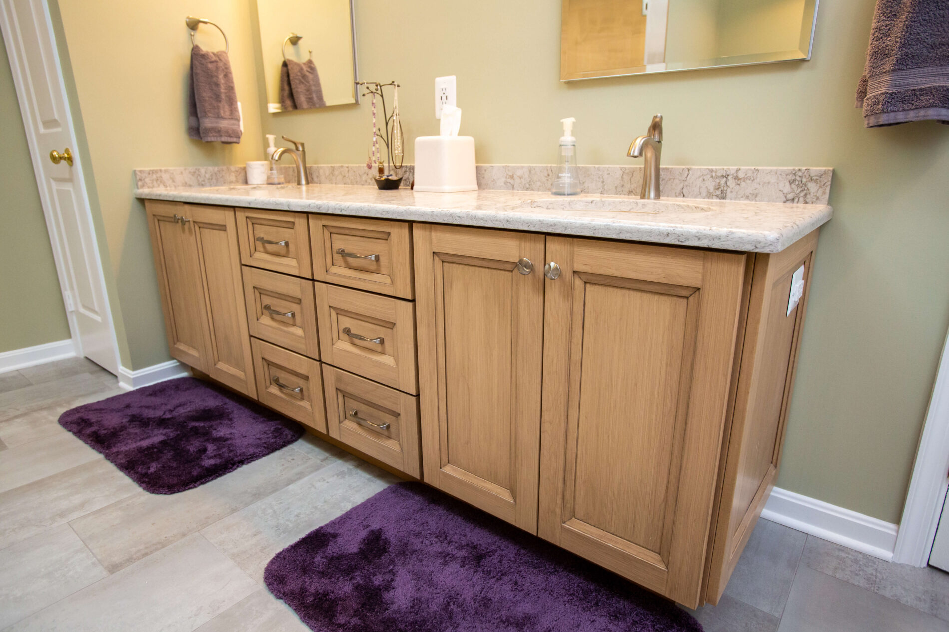 Double bathroom vanity with light wood cabinets, marble countertop, two sinks, wall-mounted faucets, and two purple bath mats on a tiled floor.