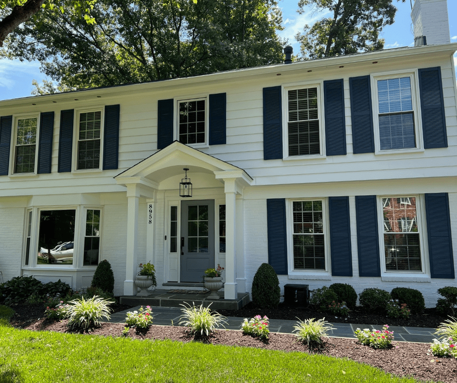 Two-story white house with dark blue shutters, centered front door, six windows, and landscaped garden with shrubs and flowers in front.