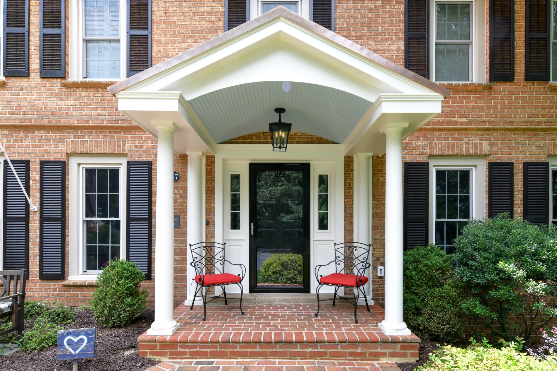 A brick house front porch with two black chairs with red cushions, a black door, white columns, black shutters, and a heart-shaped yard sign on the left.