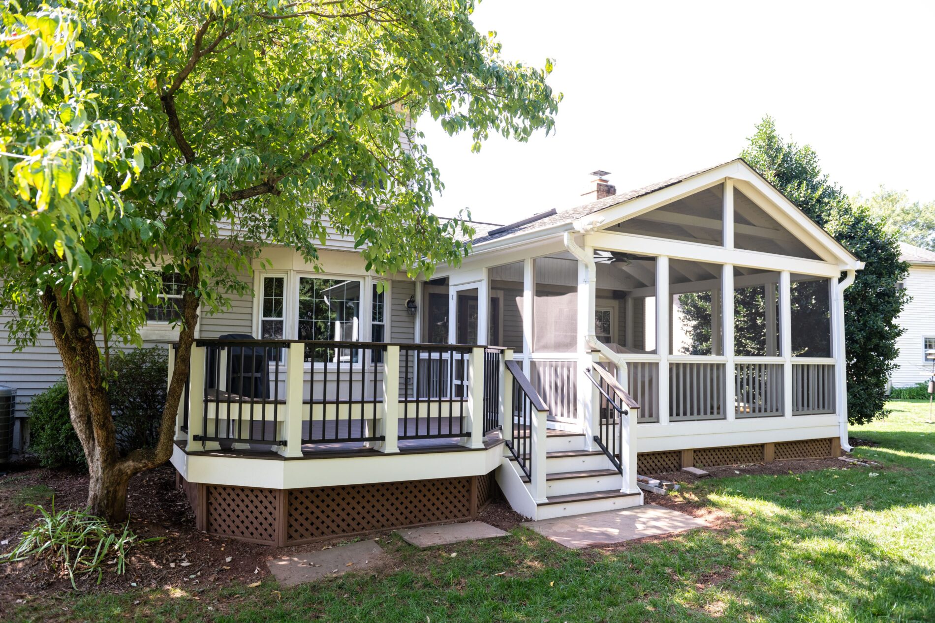 A screened porch with steps and a small deck attached to a suburban house, surrounded by a grassy yard and trees.
