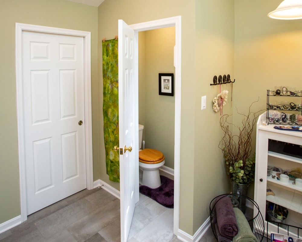 Bathroom with light green walls, a white closet door, a partially open door to a toilet with a wooden seat, green shower curtain, and shelves with rolls and decor items—an inviting space that highlights functional home design.