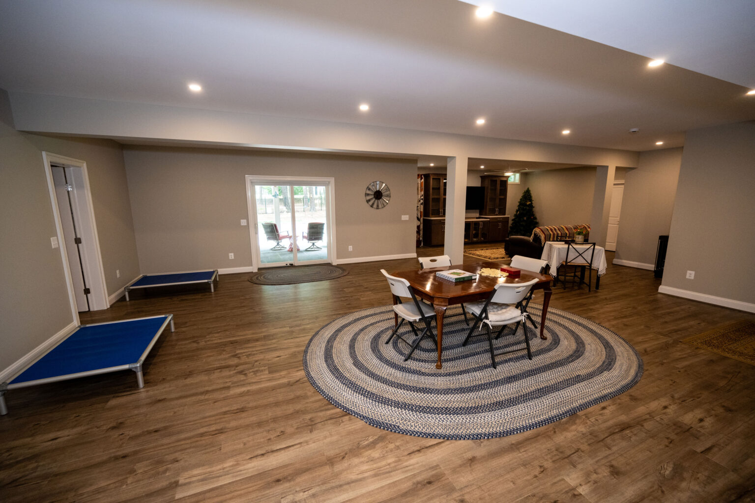 Spacious room with wood flooring, a round rug, table and chairs in the center, two blue pet beds, and sliding glass doors at the back.