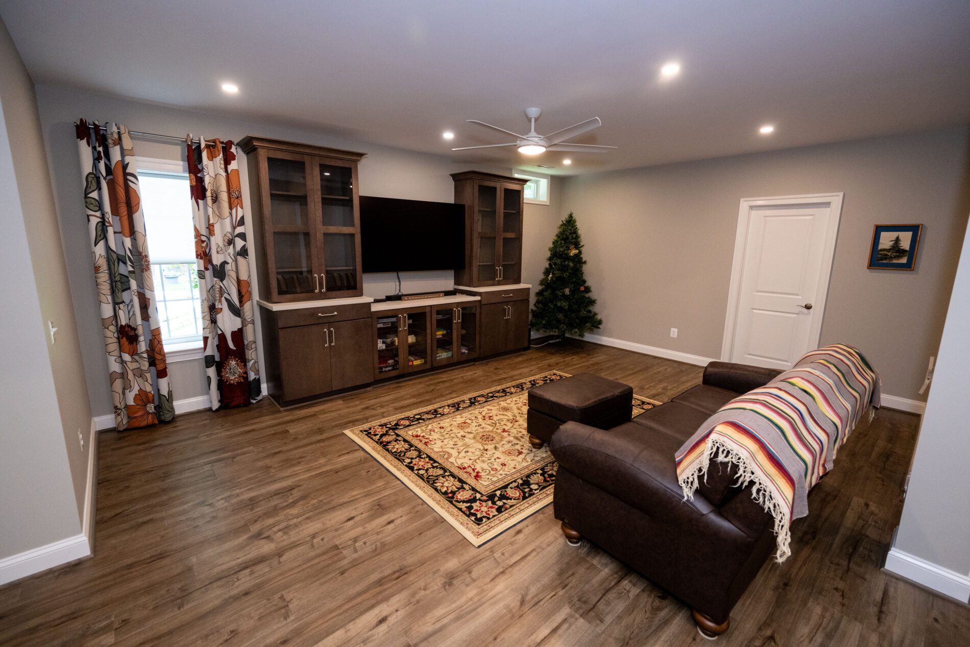 Living room with wood flooring, a rug, brown sofa, ottoman, TV on a cabinet, glass display shelves, a Christmas tree, and curtains with floral prints. Ceiling fan and recessed lights overhead.