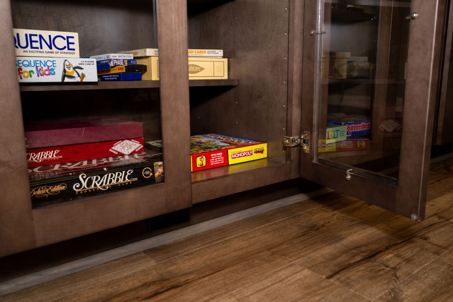 Several board games, including Scrabble, Monopoly, and Sequence, are stored on shelves inside a wooden cabinet with glass doors, one door open, on a wooden floor.