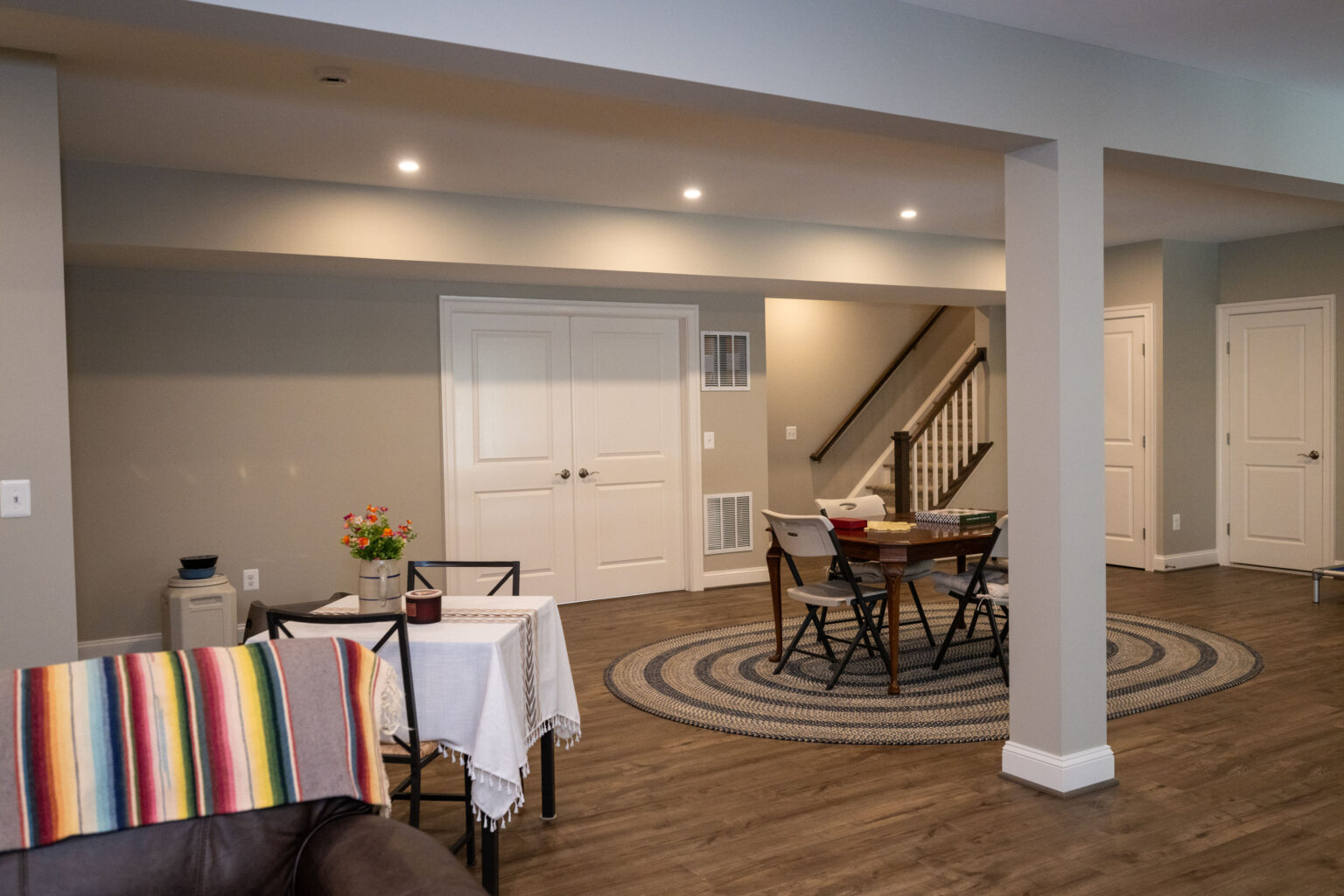 Modern basement with wood flooring, round rug, dining table with chairs, small covered table, and staircase in the background. Neutral wall colors and recessed lighting.