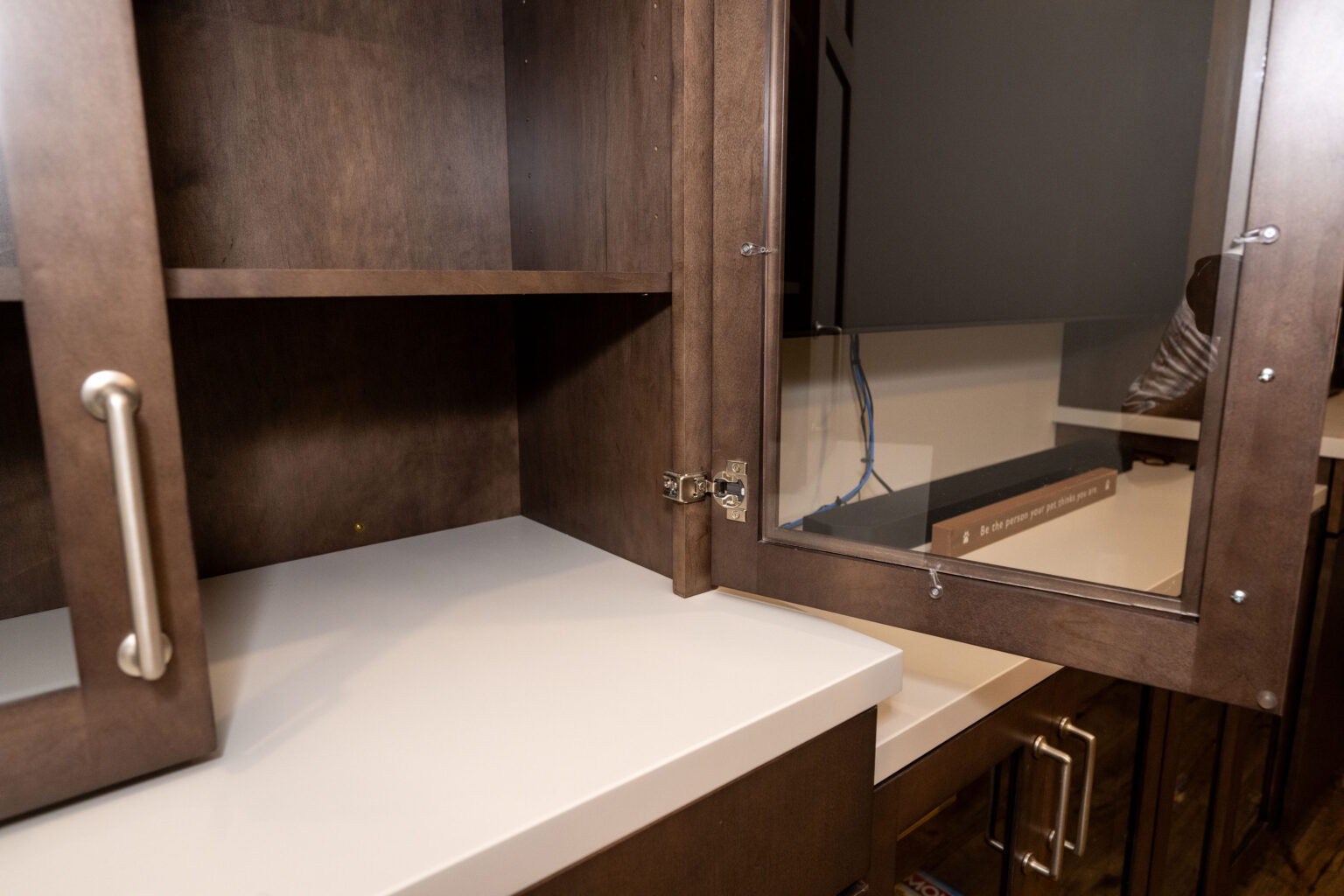 Open brown cabinet with a mirrored door, white countertop, and silver handles, showing empty shelves and part of a room reflected in the mirror.