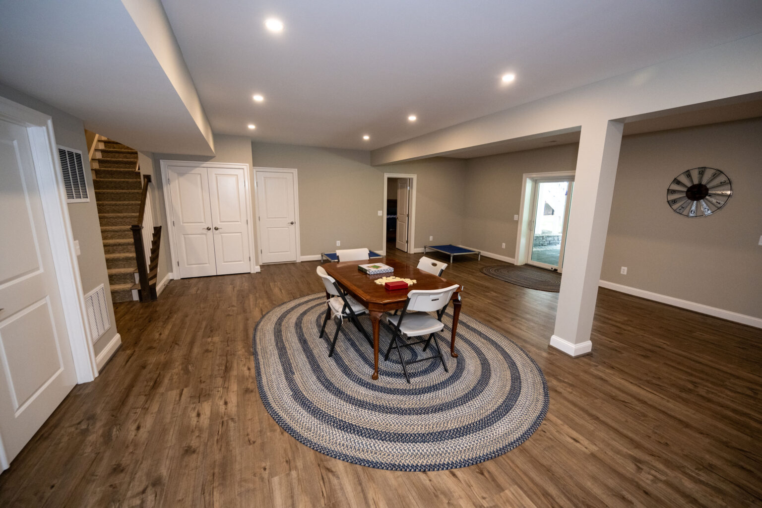 Spacious basement room with wood floors, a round rug, a table with four chairs, and a wall clock. Stairs and two white doors are on the left, with sliding doors at the back.