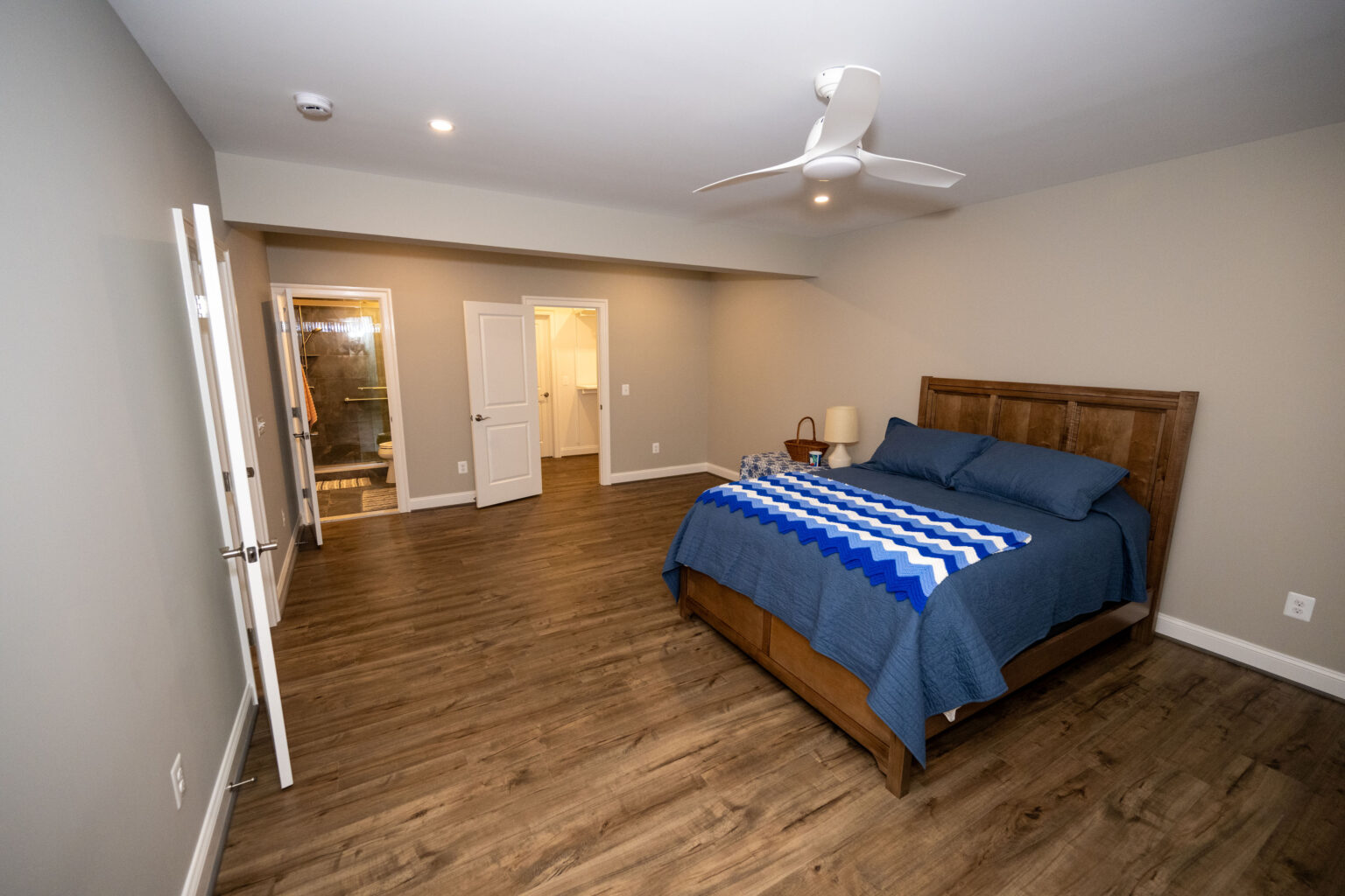 Spacious bedroom with wooden floor, a bed with blue bedding, a ceiling fan, two open doors, and neutral-colored walls.