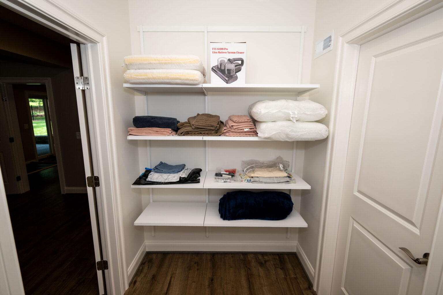 A small closet with four white shelves holding folded towels, blankets, bedding, and a vacuum box, with doors on either side and wooden floor.