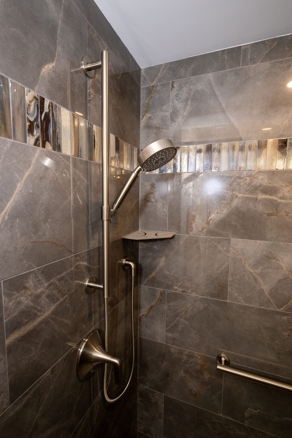 Modern shower with gray marble tile, a mounted adjustable showerhead, a small shelf, and a metal grab bar on the wall.