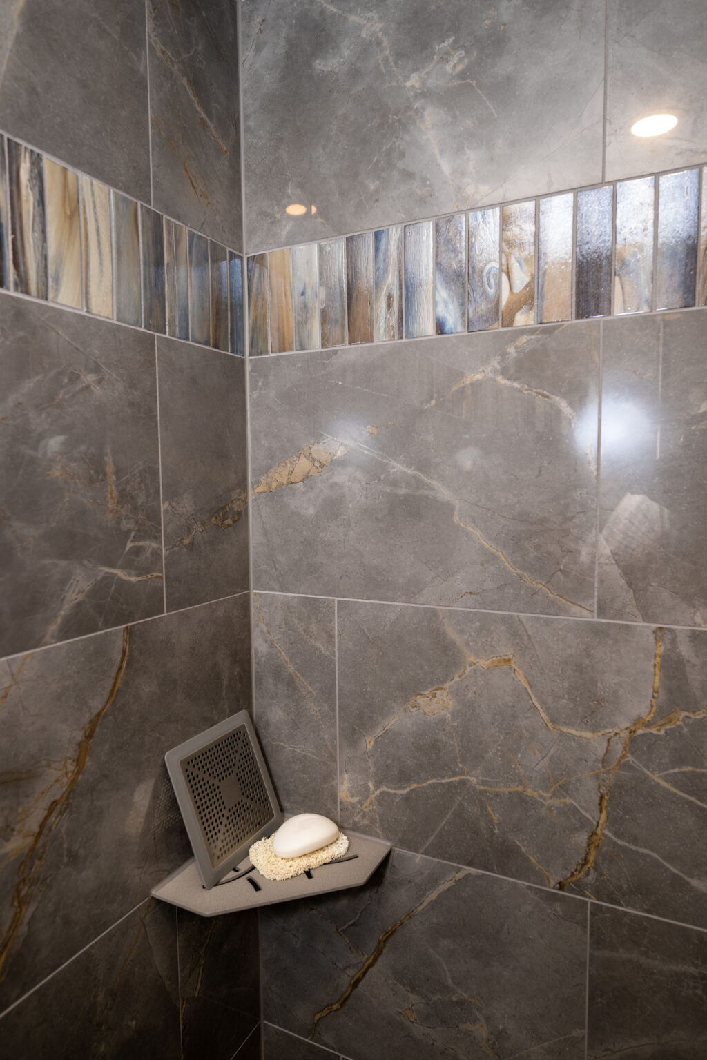 Corner of a shower with gray marble tiles, a decorative tile border, and a small triangular shelf holding a soap and a soap dish.