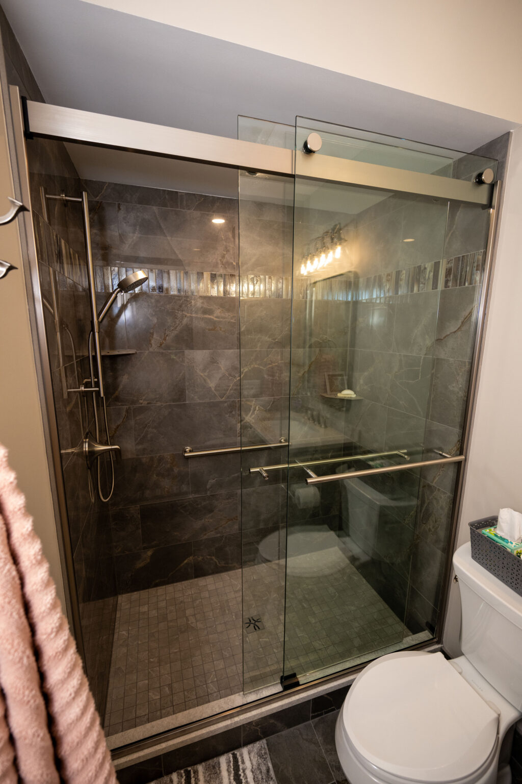 Modern bathroom with a glass-enclosed shower, wall-mounted showerhead, gray tiled walls, and a white toilet visible in the foreground.