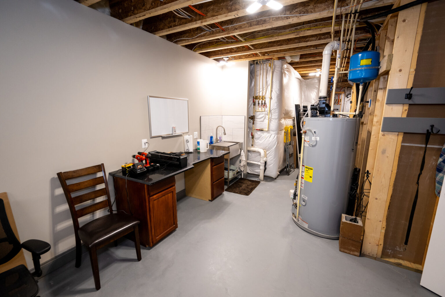 Unfinished basement with workbench, chair, water heater, exposed ceiling beams, pipes, and utility equipment against the wall.