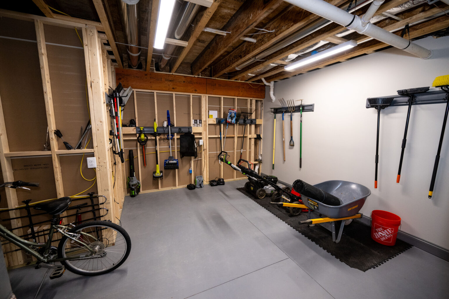 Organized garage with gardening tools, a wheelbarrow, a red bucket, a bicycle, and exposed ceiling pipes. Tools are neatly hung on wooden wall racks.