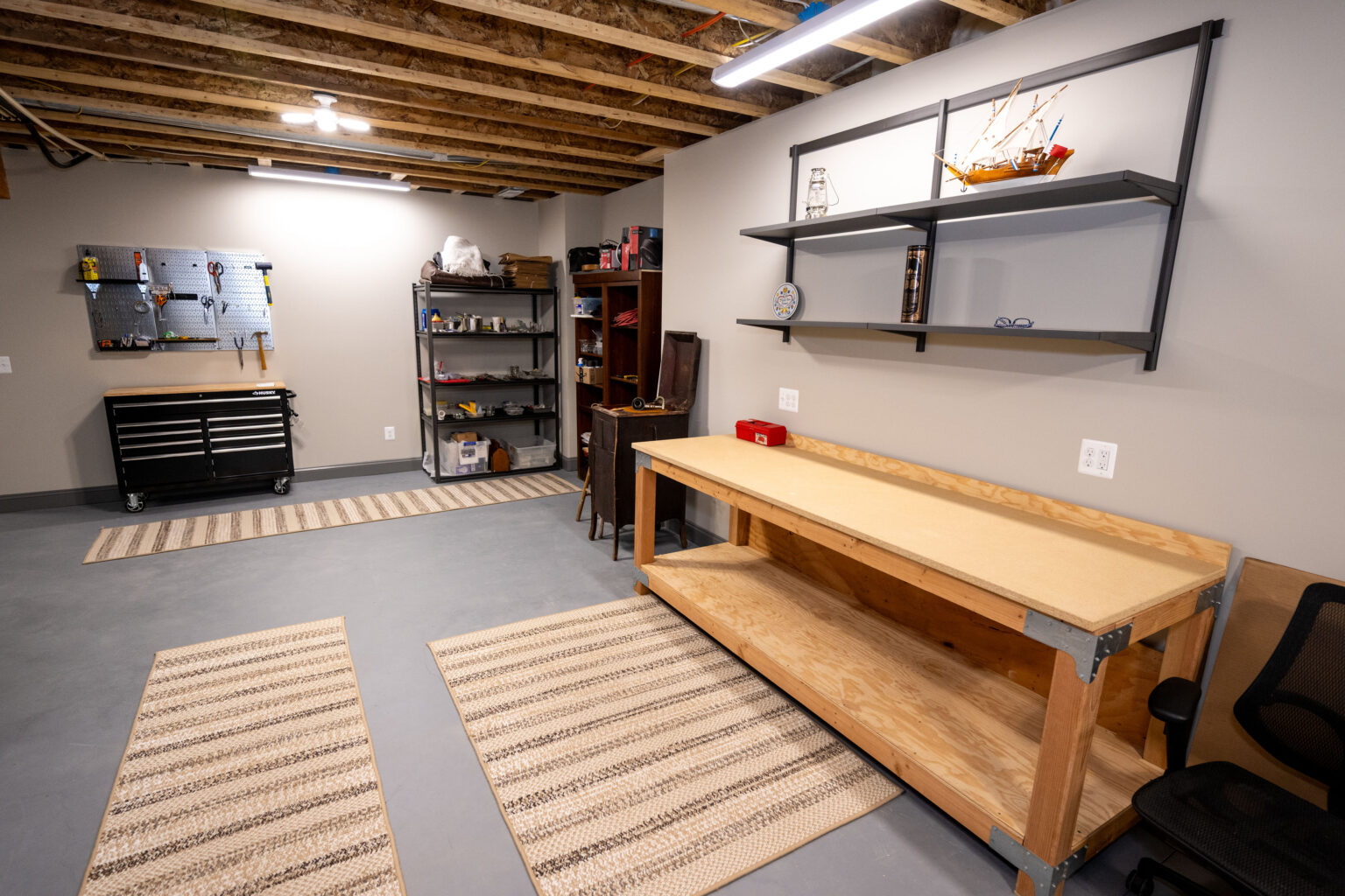 Organized basement workshop with workbench, pegboard, shelves, rugs, and exposed ceiling beams. Tools and supplies are neatly arranged; the space is clean and well-lit.