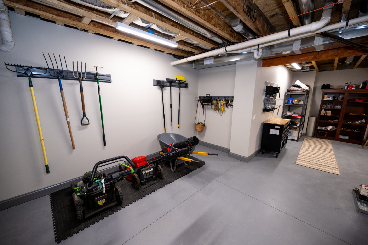 A clean basement workshop with tools and gardening equipment organized on walls, a wheelbarrow and power tools on the floor, and shelves with supplies in the background.