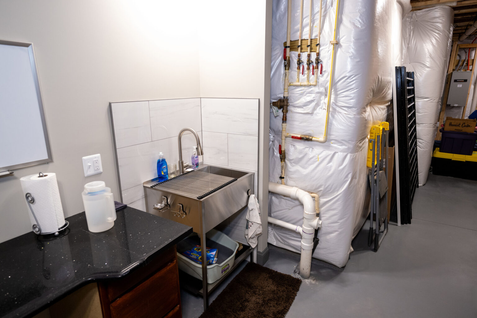 A utility sink with cleaning supplies is next to a black counter in a basement laundry or utility room. Insulated pipes and storage bins are visible along the wall.