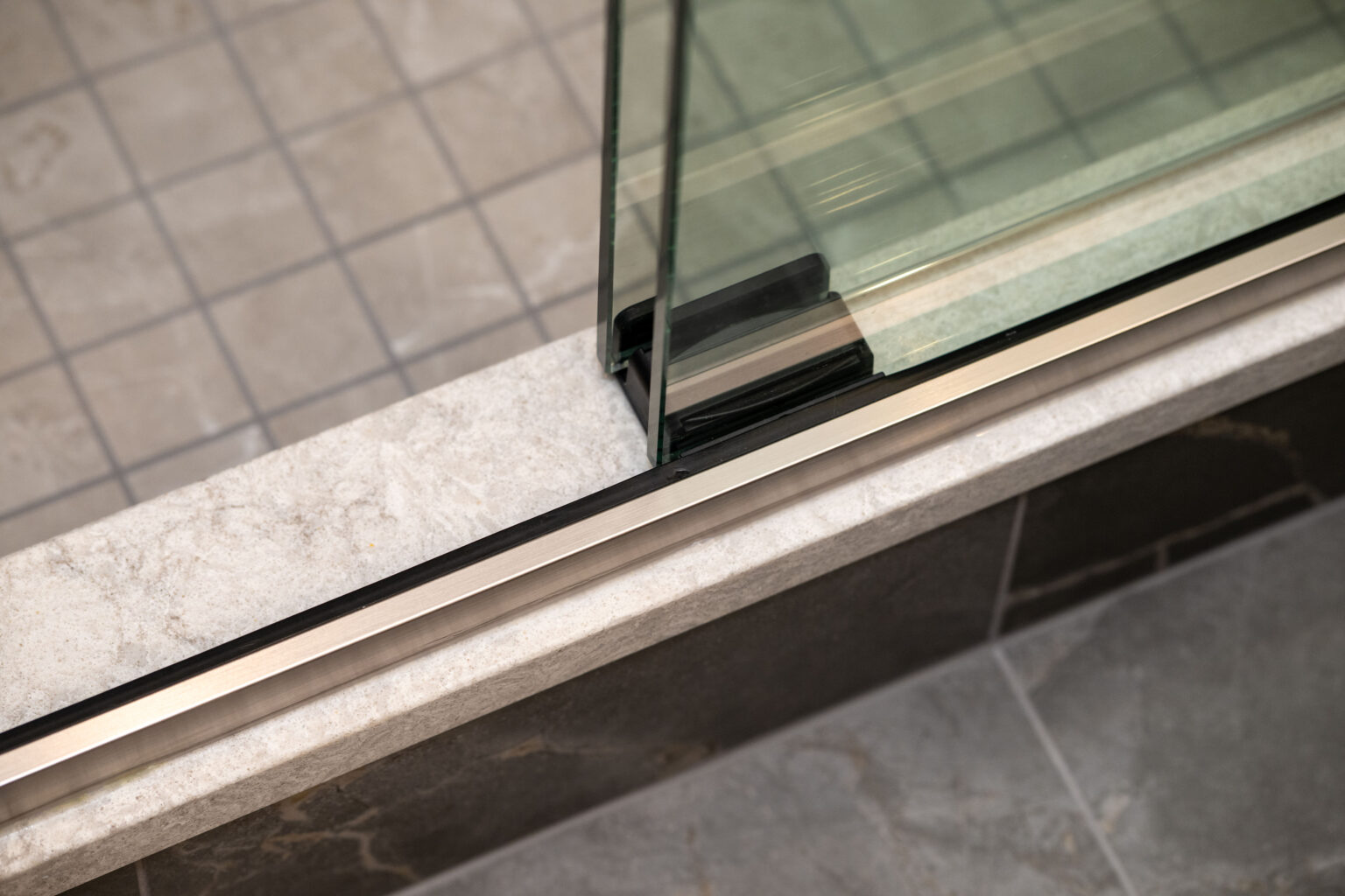 Close-up of a glass shower door with a metal frame set on a tiled ledge, showing the corner detail between the glass, metal, and tile surfaces.