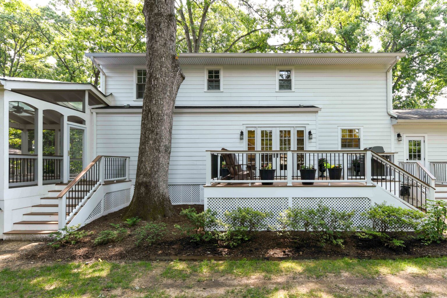 White two-story house with a screened porch, deck, and stairs, surrounded by trees and shrubs, on a sunny day.