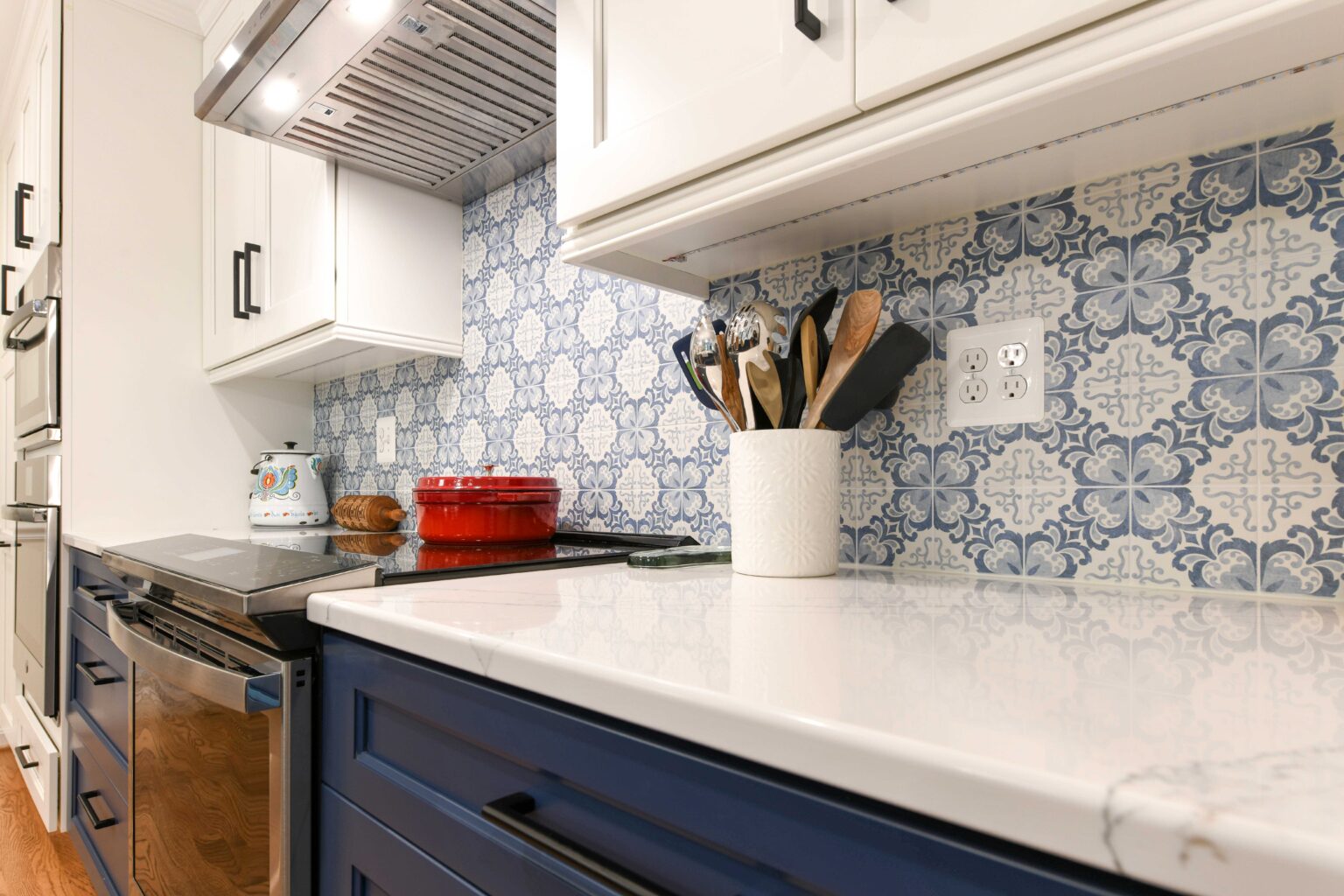 Modern kitchen with blue and white patterned backsplash, white countertop, blue cabinets, utensils in a holder, and a red pot on the stove.
