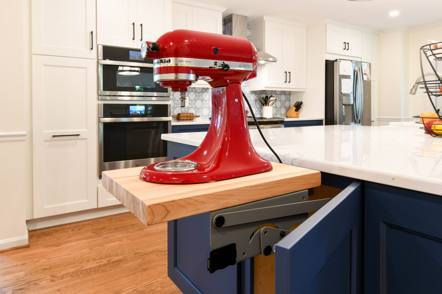 A red stand mixer sits on a pull-out wooden shelf built into a blue kitchen cabinet in a modern kitchen.