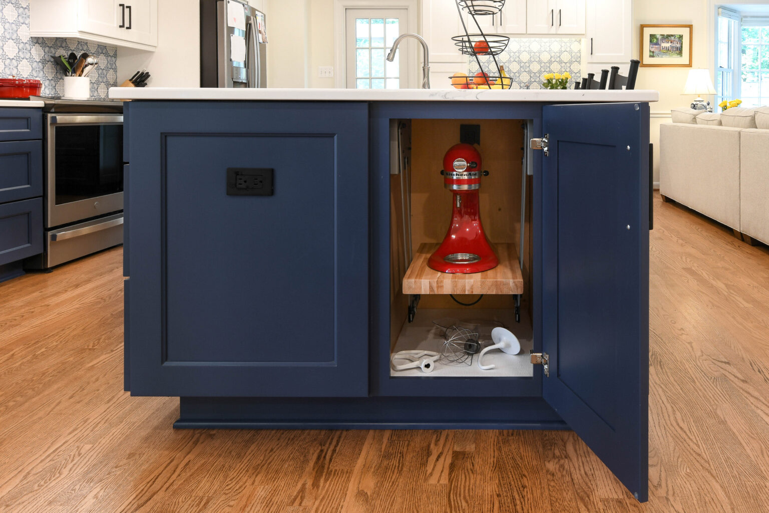 A red stand mixer sits on a pull-out shelf inside a blue kitchen island cabinet with the door open. Baking attachments are on the bottom shelf below.