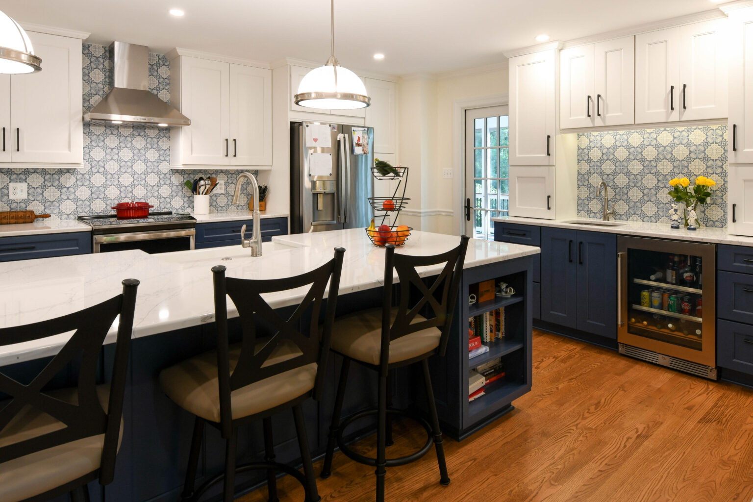 Modern kitchen with a large island, three chairs, blue and white cabinets, patterned tile backsplash, stainless steel appliances, and wooden floor.