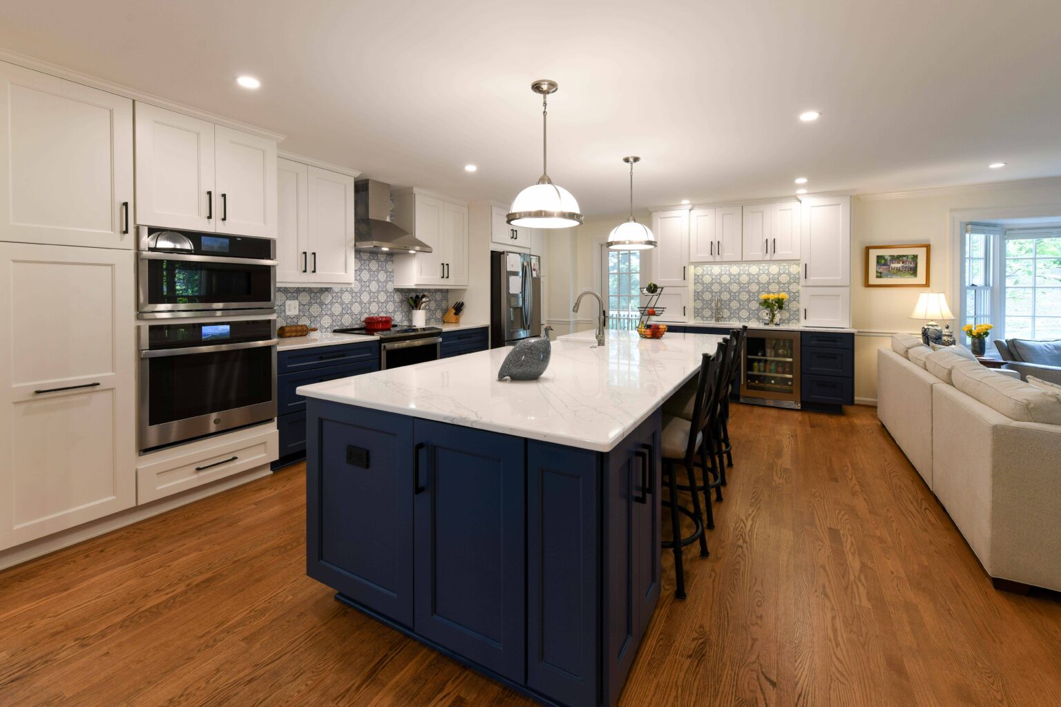 Modern kitchen with white cabinets, navy blue island, marble countertops, stainless steel appliances, wood floor, tile backsplash, and built-in oven. Living area visible on the right.