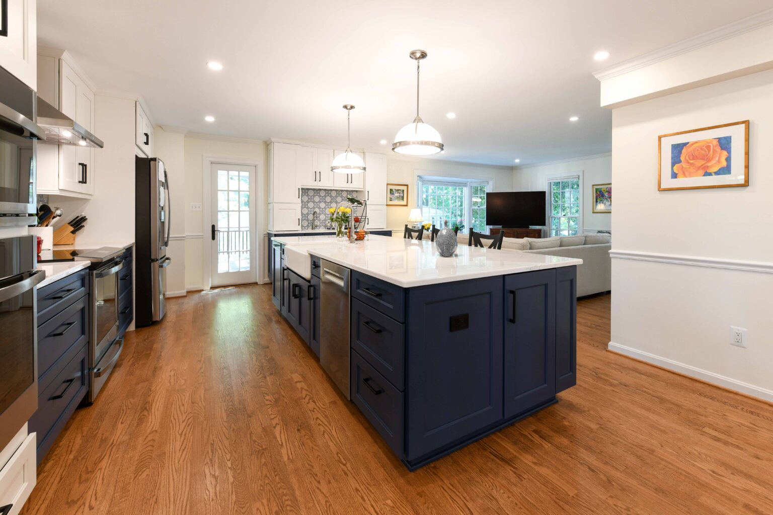 Modern kitchen with blue island, white cabinets, wood flooring, stainless steel appliances, pendant lights, and adjoining living room with TV and couches in the background.