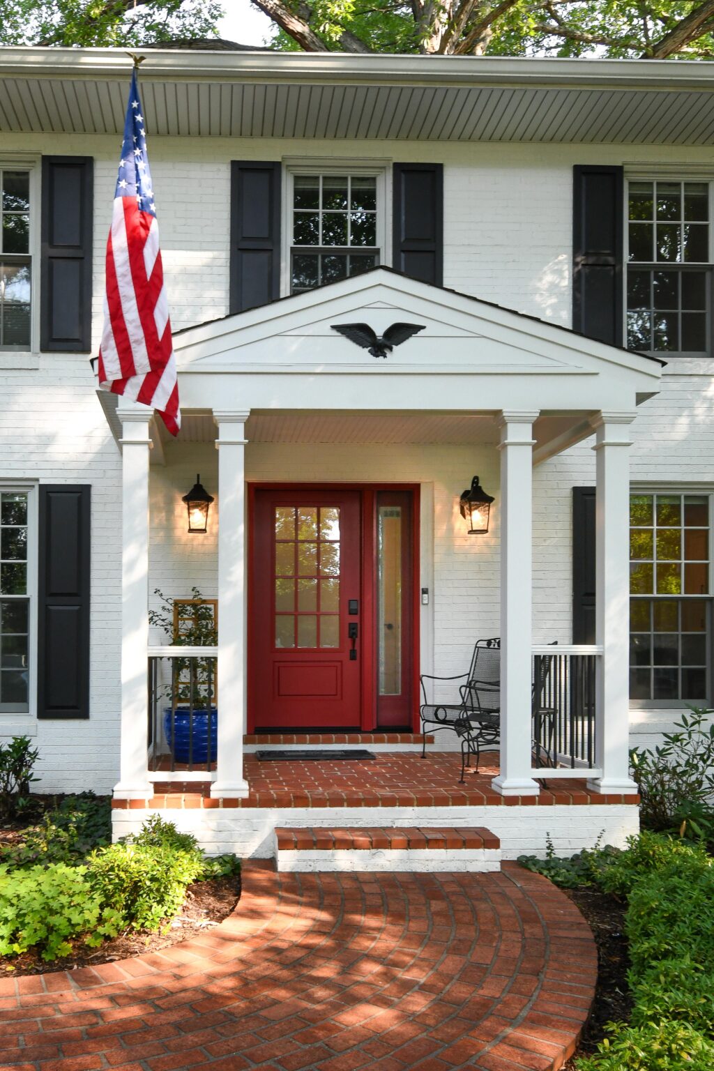 Two-story white brick house with black shutters, a red front door, a small porch with columns, an American flag, and a curved brick pathway leading to the entrance.