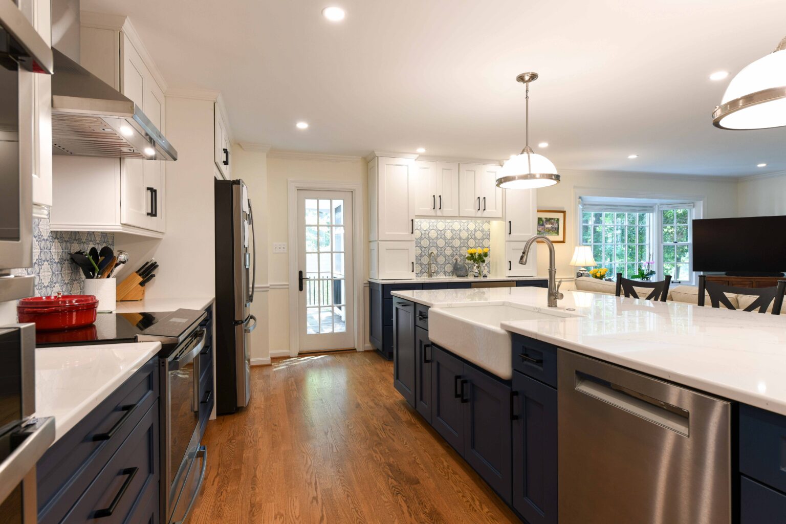 Modern kitchen with white and navy cabinets, stainless steel appliances, farmhouse sink, pendant lights, hardwood floor, and large windows providing natural light.