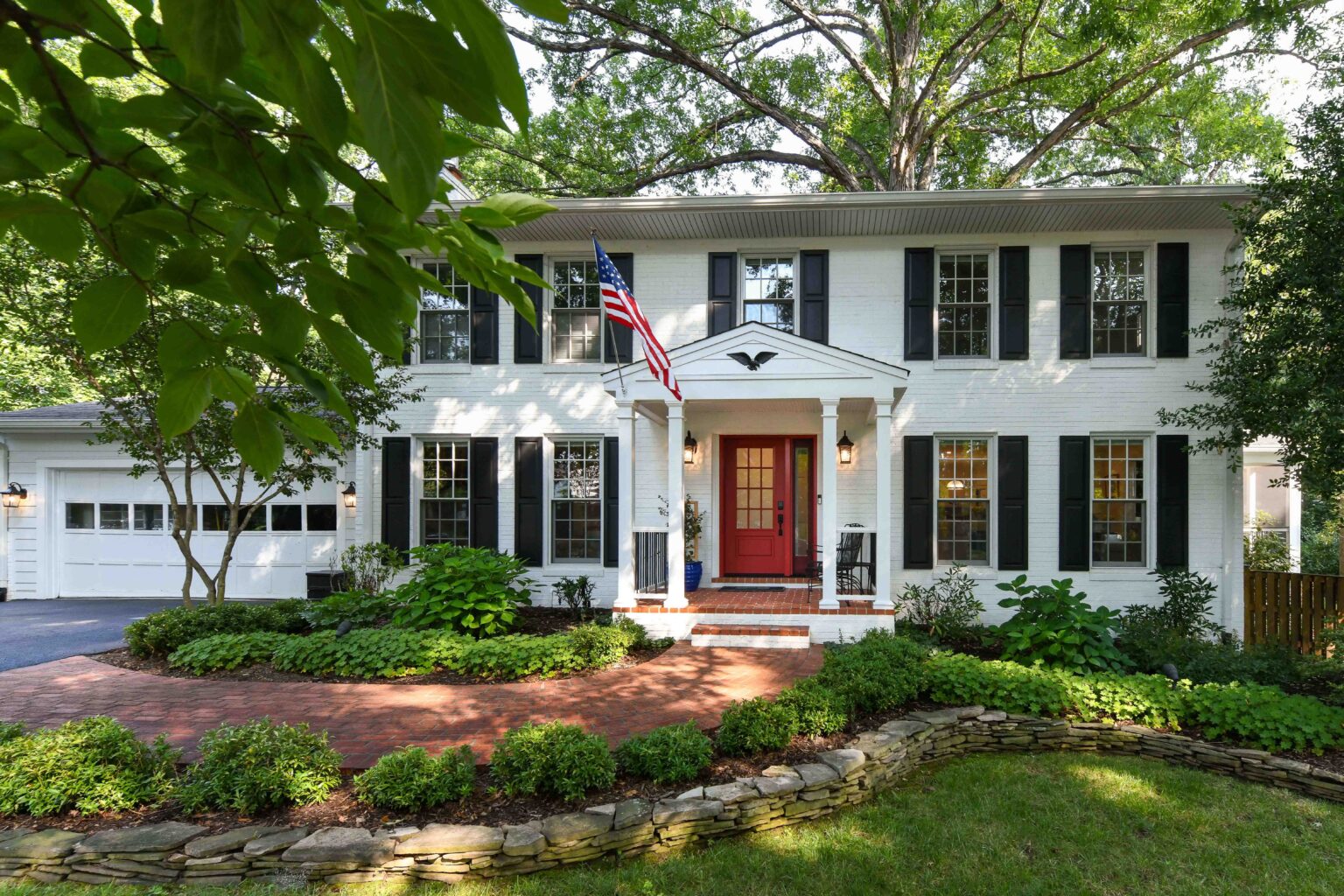 Two-story white brick house with black shutters, a red front door, American flag, attached garage, landscaped yard, and brick walkway, surrounded by trees and greenery.