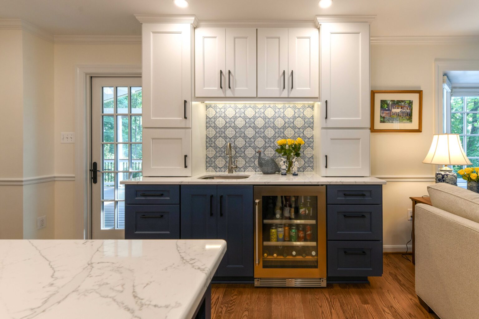 Modern kitchen area with blue and white cabinets, a patterned tile backsplash, a small sink, built-in beverage fridge, and decorative flowers on a marble countertop.