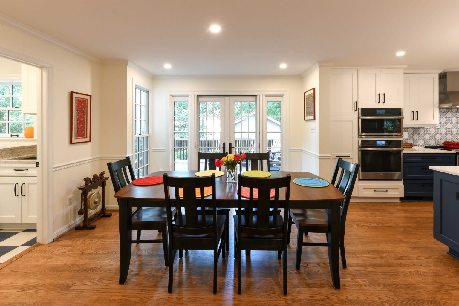 Dining area with six black chairs around a wooden table, colorful plates, bright kitchen, white cabinets, and large windows letting in natural light.