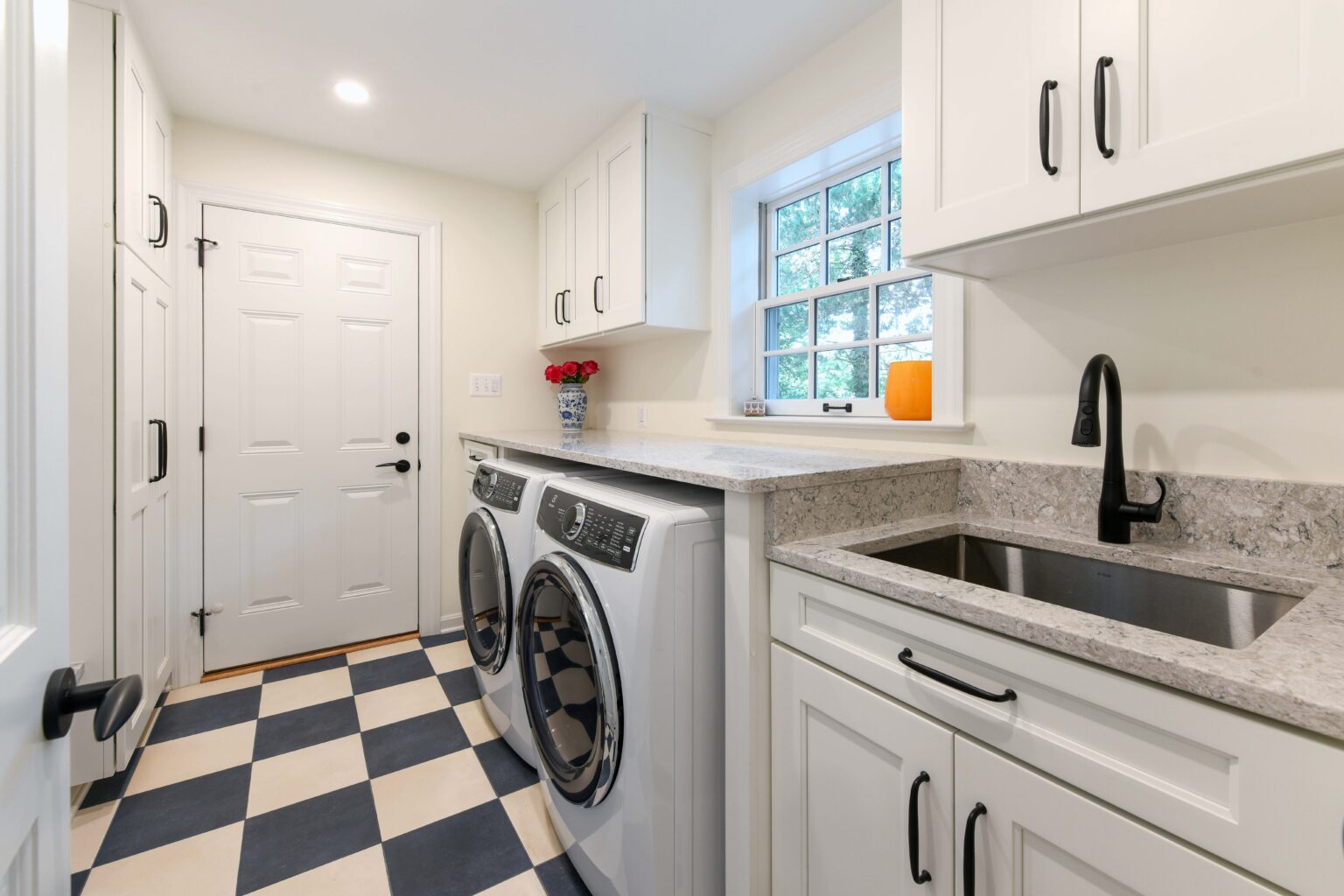 Laundry room with checkered tile floor, white cabinets, washer and dryer, stainless steel sink, and window; a vase of red flowers and an orange candle sit on the counter.