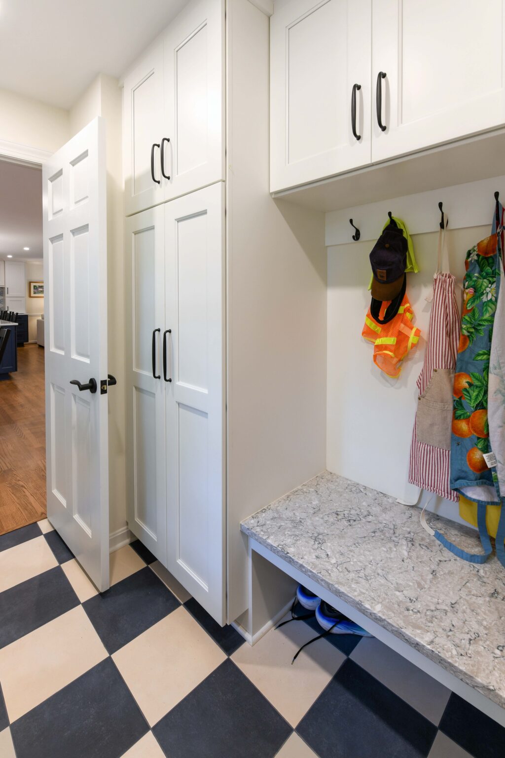 Mudroom with white cabinets, black handles, a marble bench, black hooks holding hats and aprons, and black-and-white checkered tile floor.