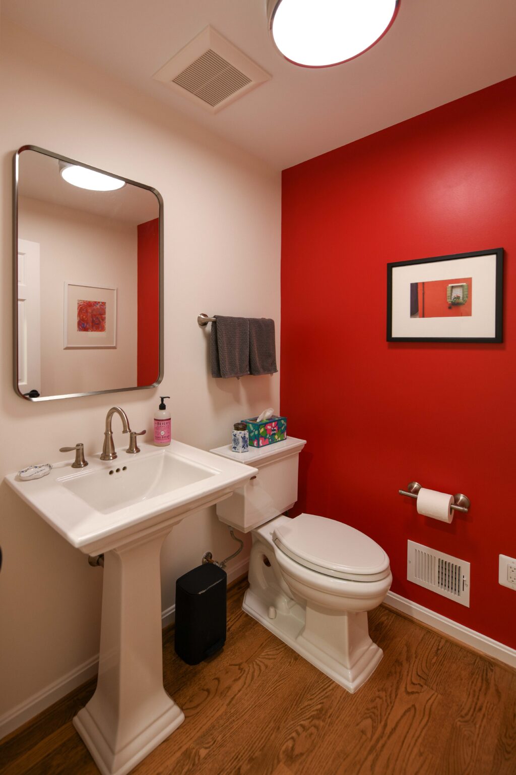 A bathroom with a pedestal sink, toilet, towel rack, mirror, red accent wall, artwork, wood floor, and toiletries on the toilet tank.