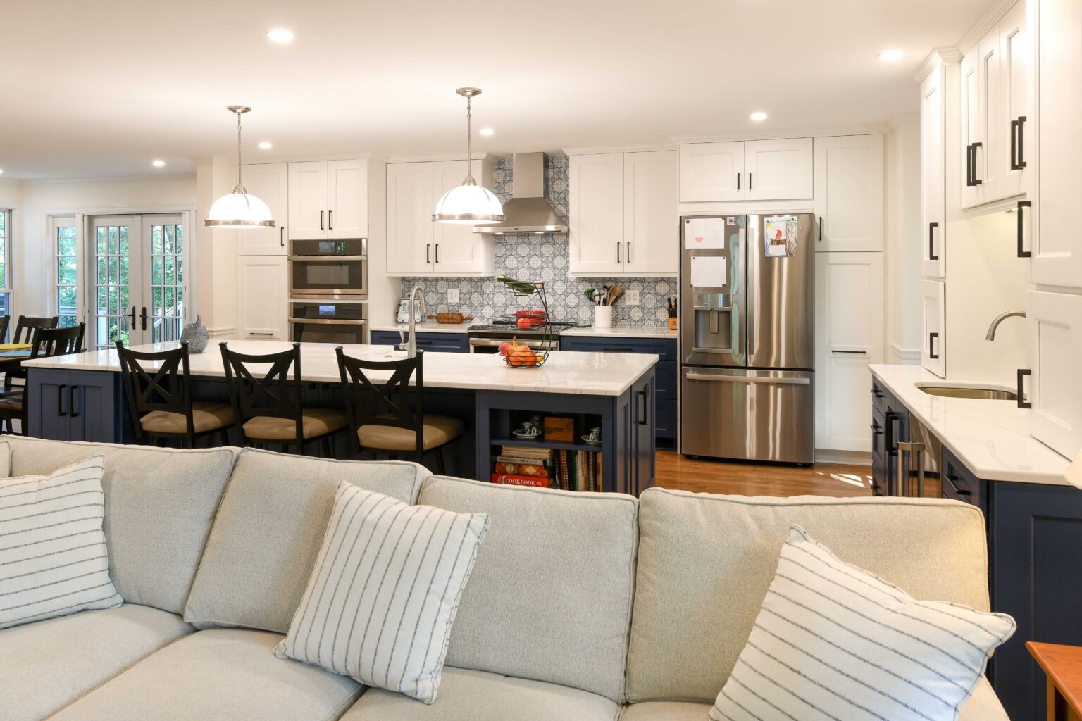 Modern kitchen with white cabinets, stainless steel appliances, a blue island with barstools, pendant lights, and a sofa with striped pillows in the foreground.