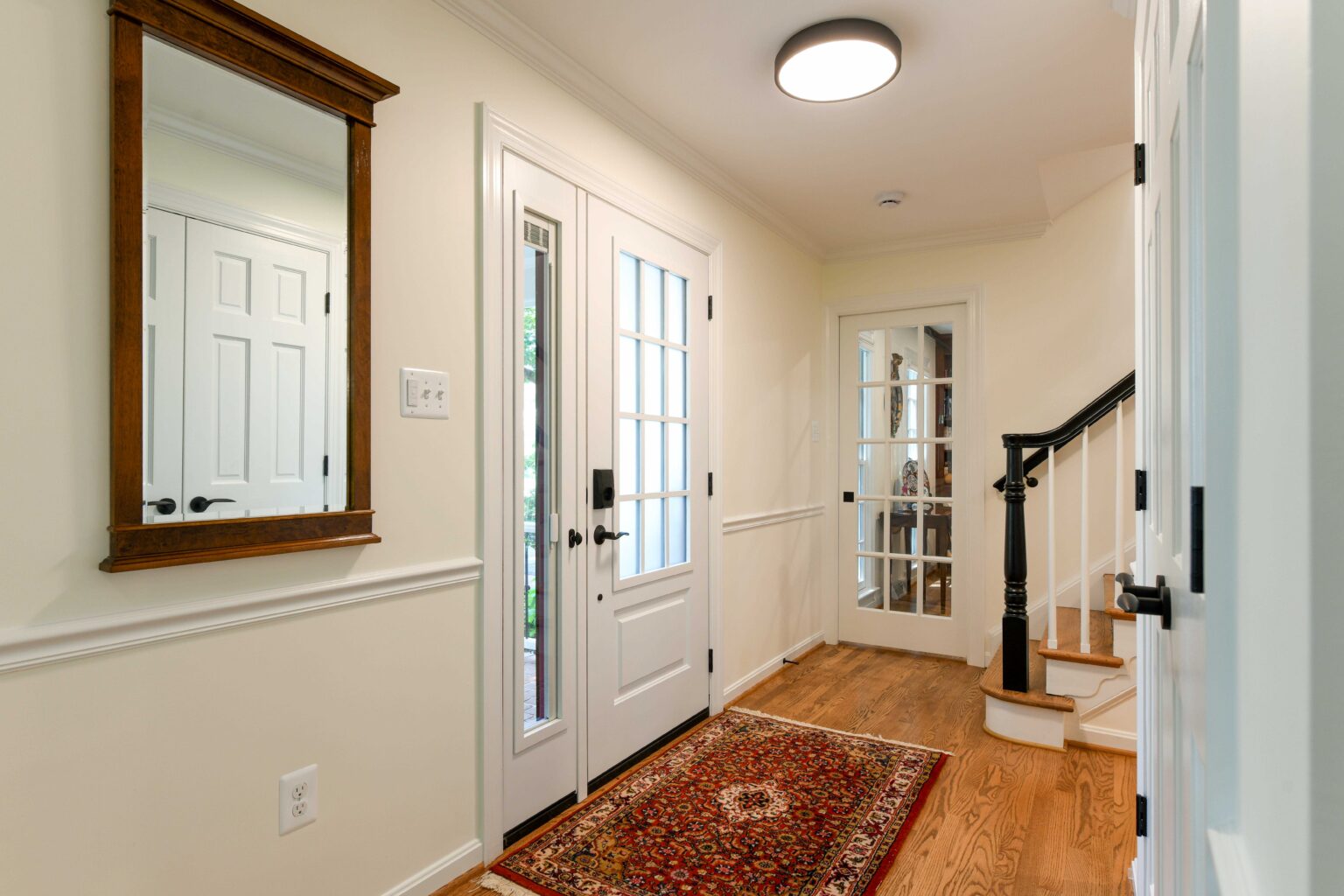 Bright entryway with white walls, a wooden floor, a patterned rug, a mirror, a white front door with glass panels, and a staircase with a black railing.