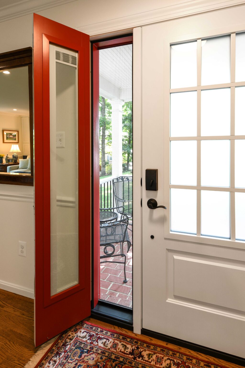 A white front door with a black handle is open, and a red storm door is ajar, revealing a brick porch with a black metal chair and greenery outside.
