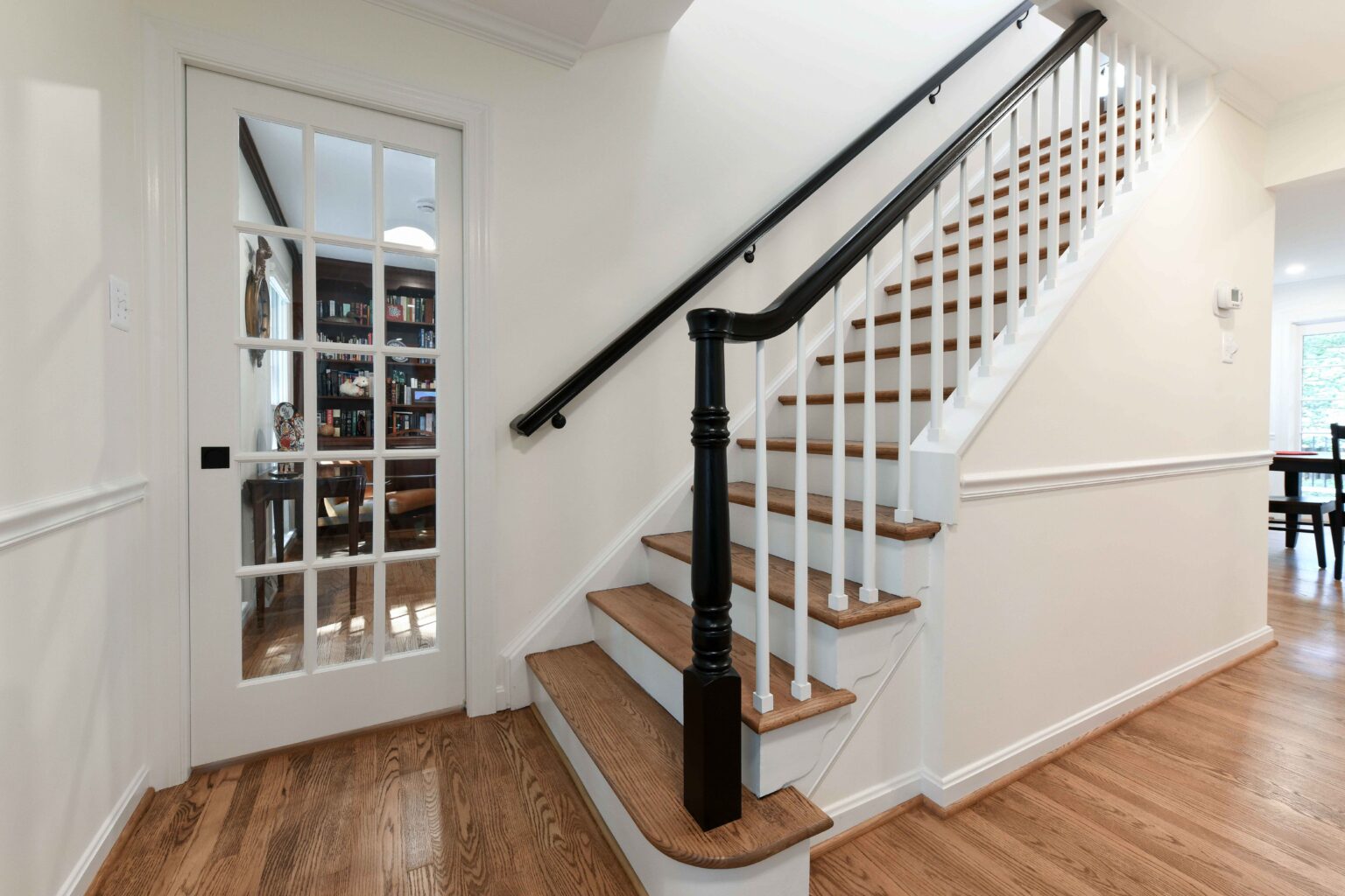 A staircase with wooden steps and white risers is next to a glass-paneled door leading to a study. The hallway has hardwood floors and white walls.